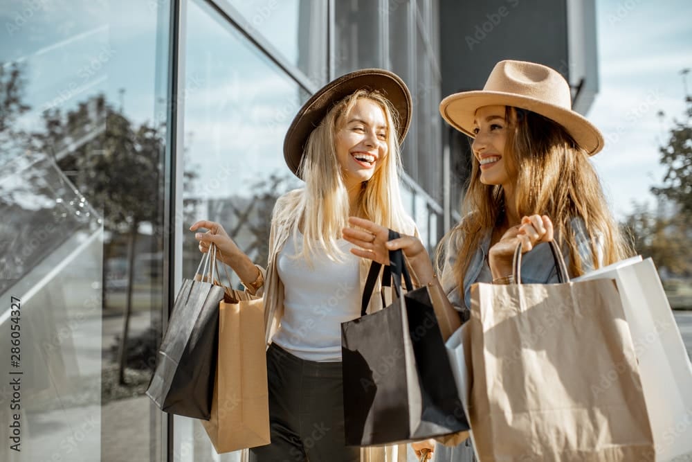 Two happy girlfriends looking on the shopwindow while standing with shopping bags near the mall because they created a budget with think and talk money